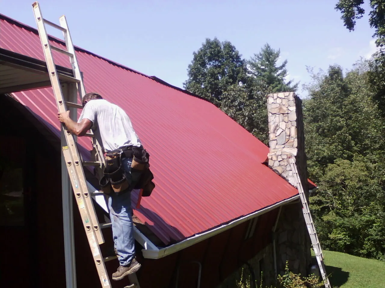 Roofer on a ladder beside a red metal roof and stone chimney — Soffit & Fascia Repair by Roofing Brothers in Coral